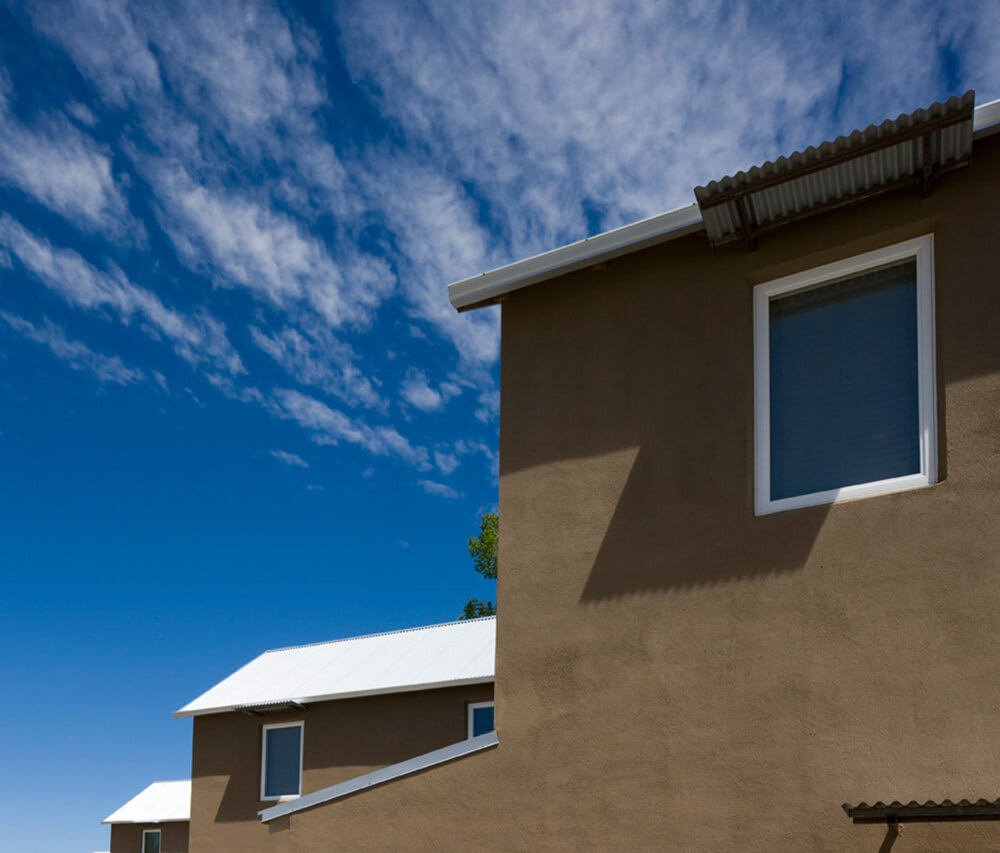 Detail of mud-toned stucco walls and metal roofs at Taos Haus against a streaked blue sky